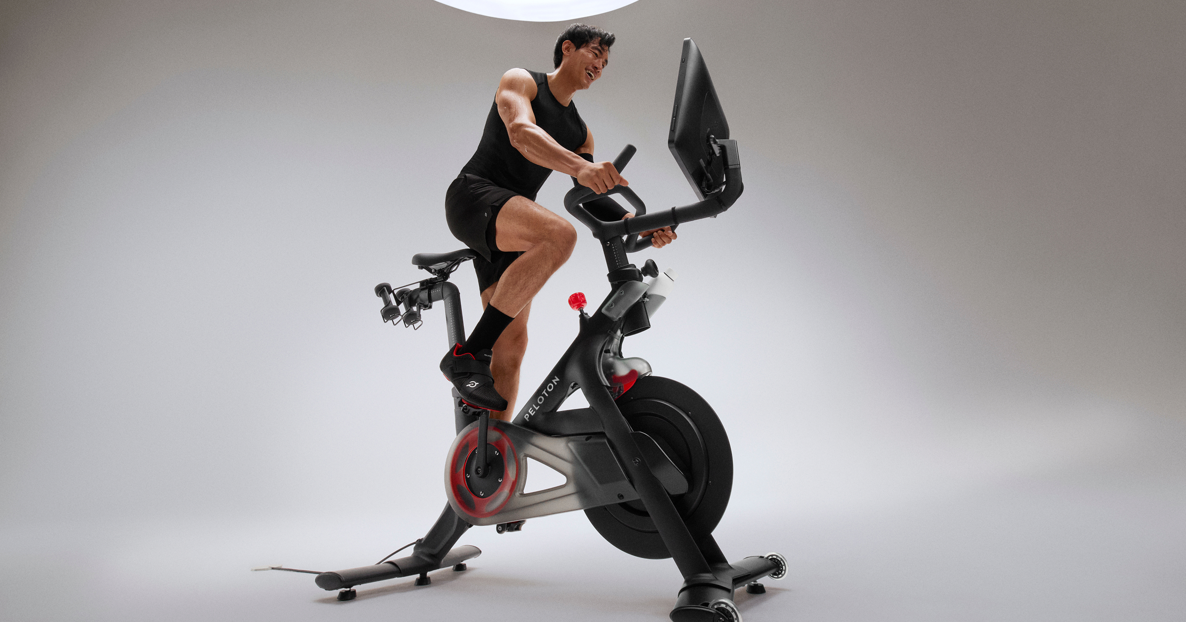 A rider pedalling hard on the Peloton bike, with the angled screen on its post and the red resistance knob below the handlebars